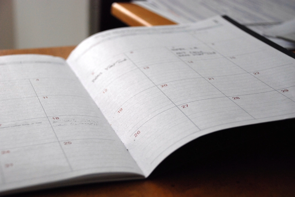 An open paper calendar on a desk, its month grid marked with handwritten notes.