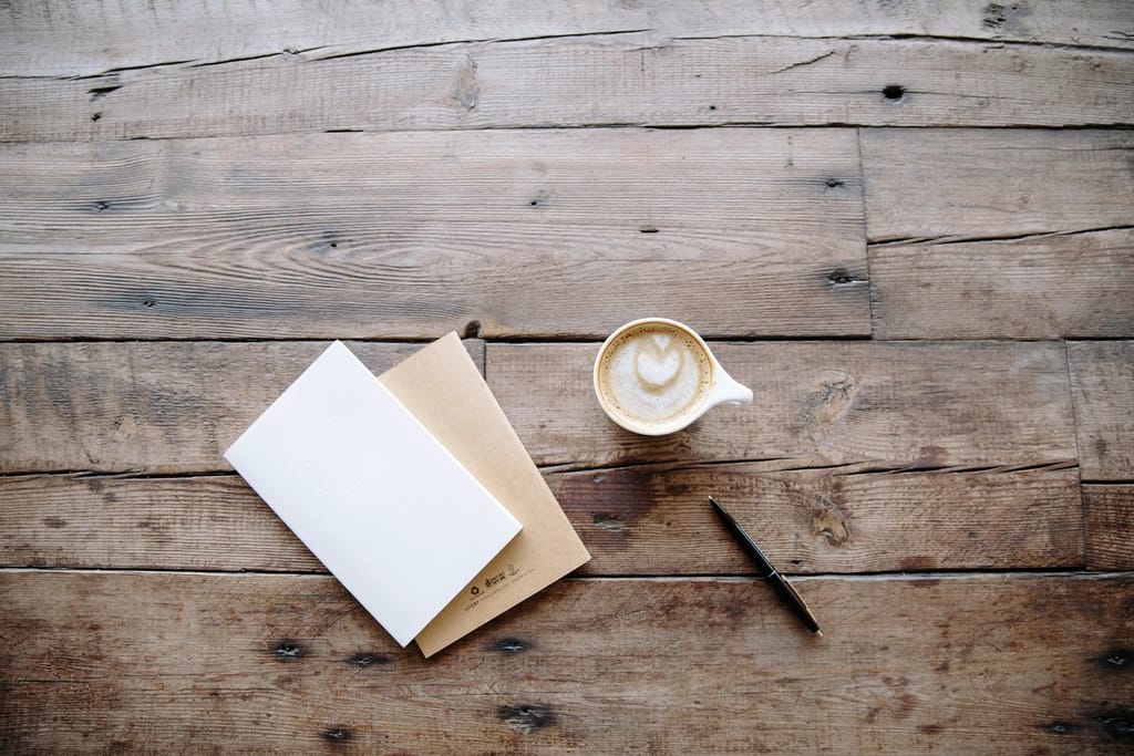 An open notebook, a pen and a cup of coffee on a bare wooden table, photographed from above.
