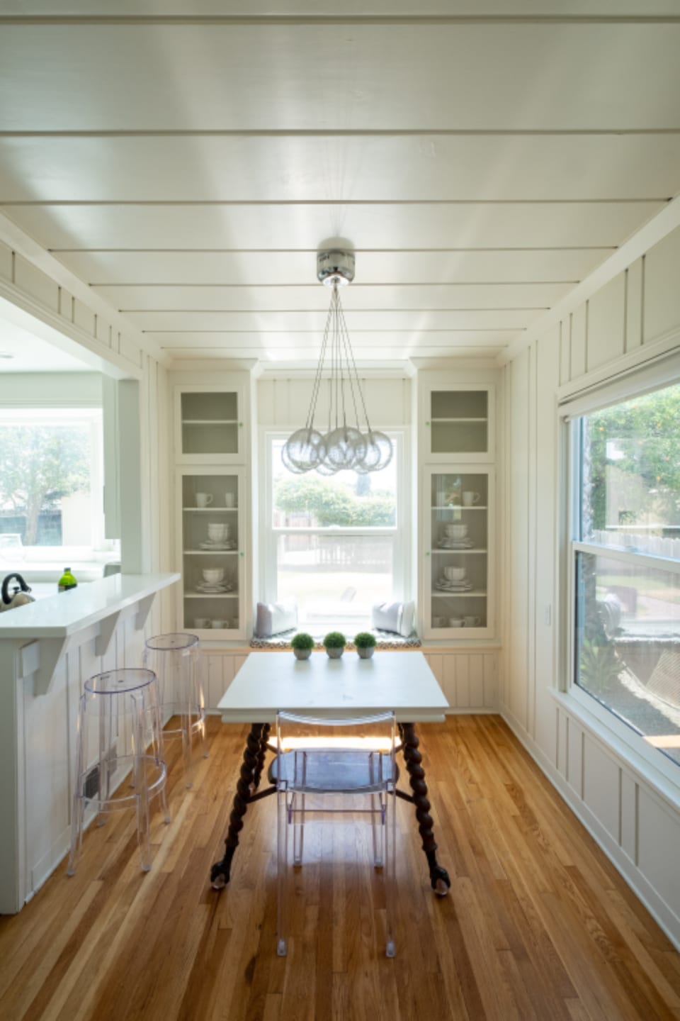 An empty dining table and chairs in a bright room, lit by a window at the far end.