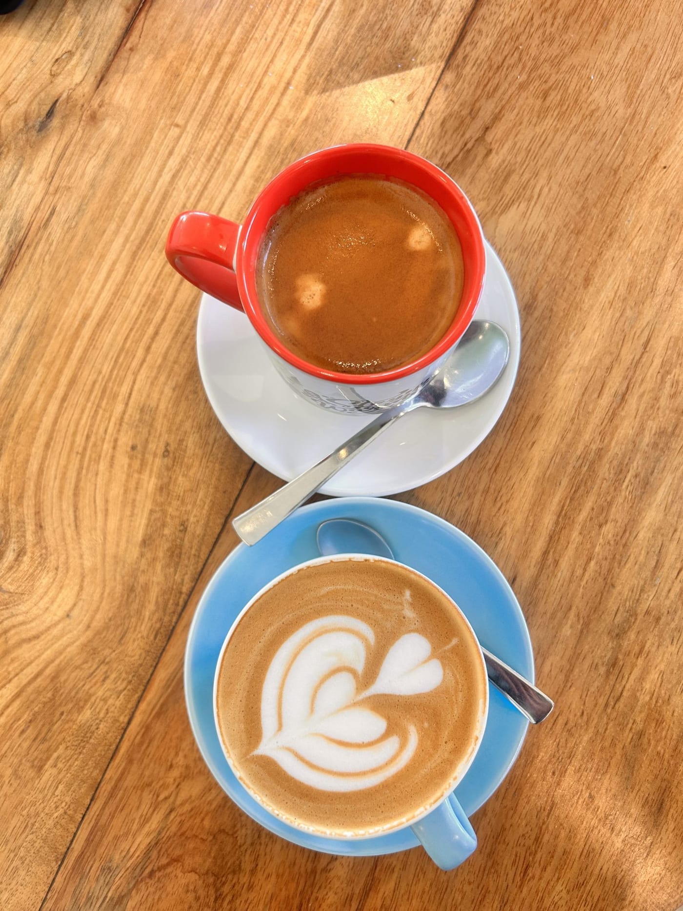 Two cups of coffee on a wooden table seen from above, one red mug and one blue cup with latte art.